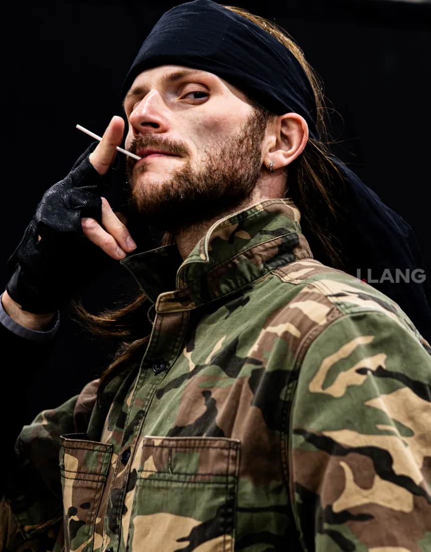 Bearded cosplayer in a black bandana and camo jacket holds a white stick to his lips