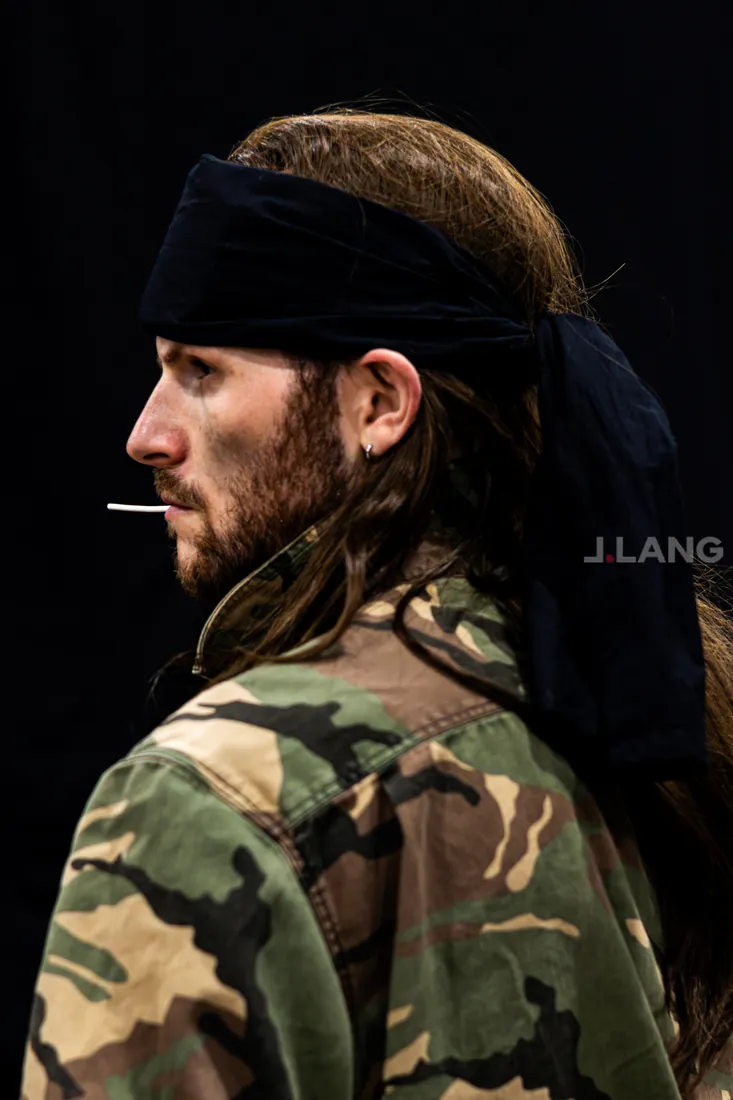Side profile of a long-haired bearded man in a black bandana and camo jacket