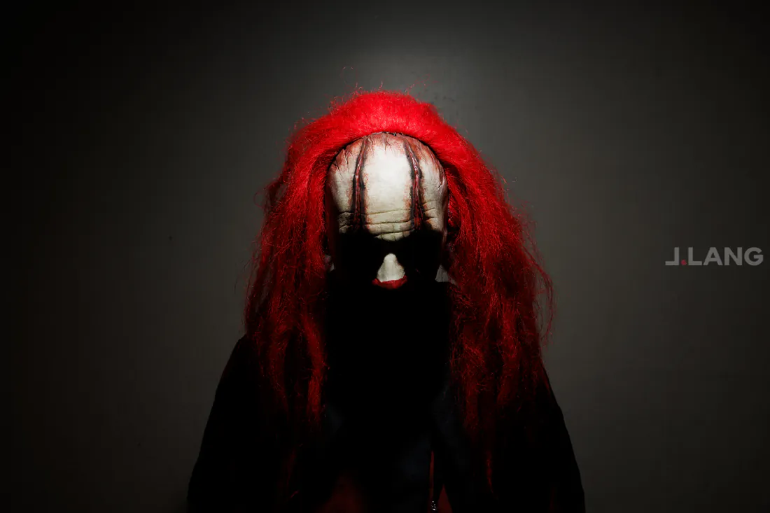 Clown in a long red wig with head bowed, only the forehead catching the light