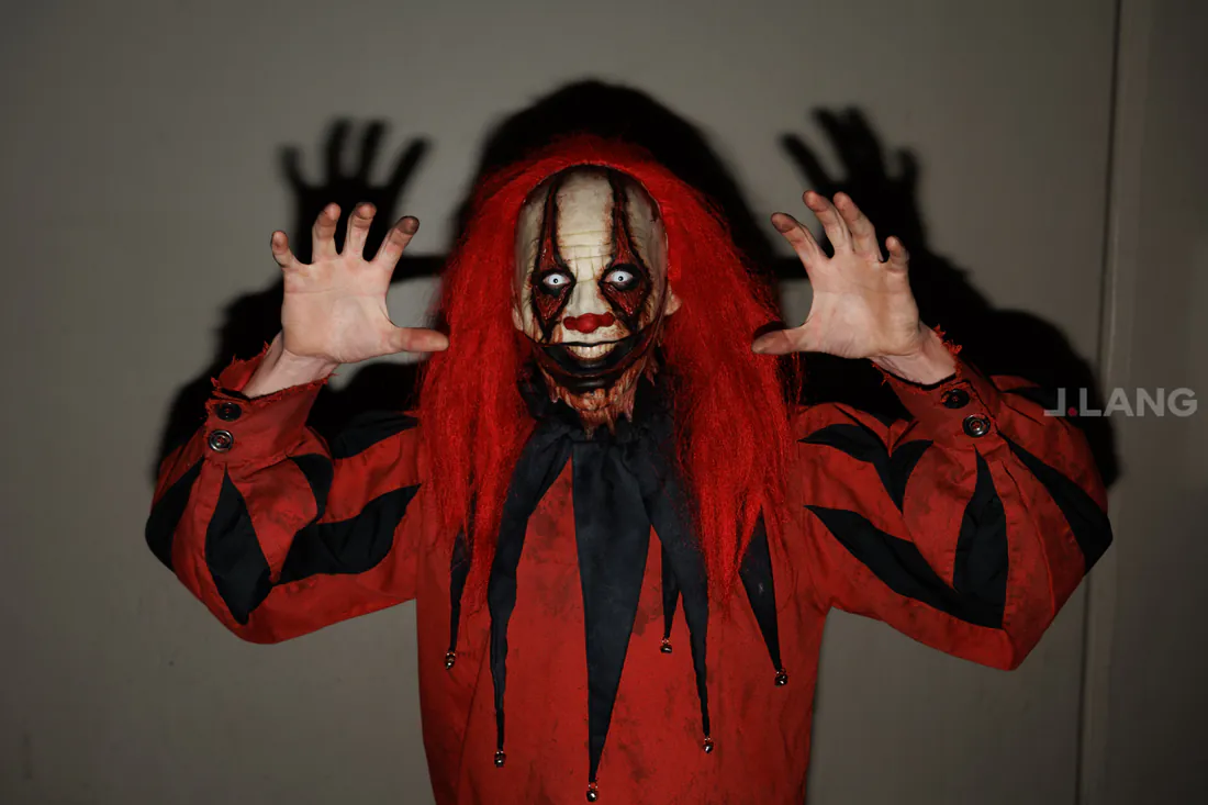 Horror clown in a red wig raises both hands as their shadows loom on the wall
