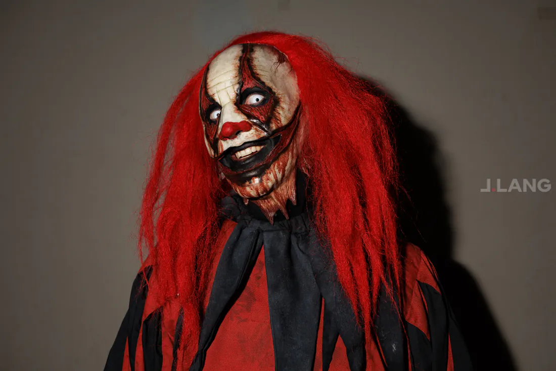 Horror clown in a red wig glances sideways with wide white eyes and a toothy grin