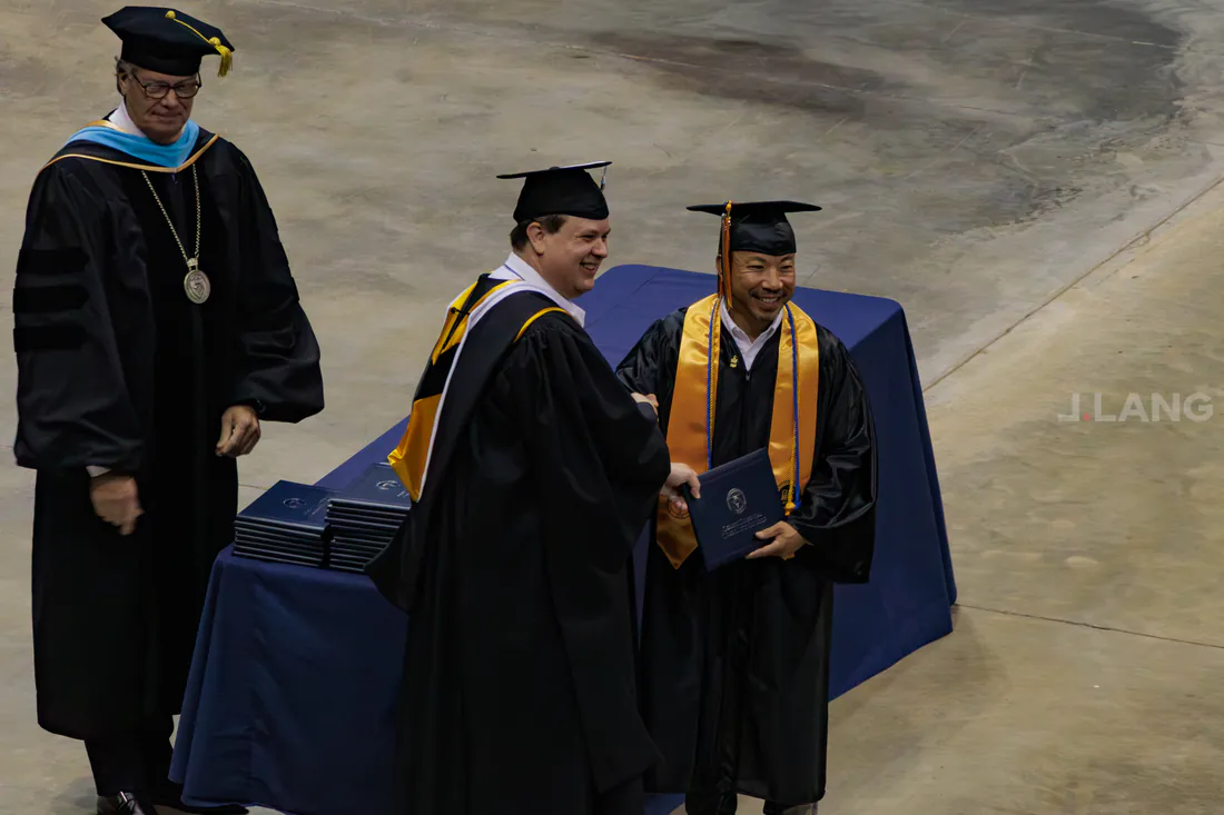 Graduate in cap and gold stole shakes hands with a robed official while holding a diploma