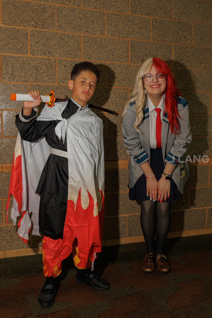 Boy in a flame-trimmed cape beside a smiling young woman in school uniform and two-tone wig