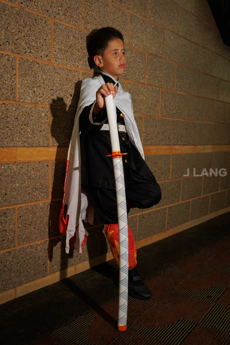Boy in a white cape leans against a wall, gripping a tall prop sword upright
