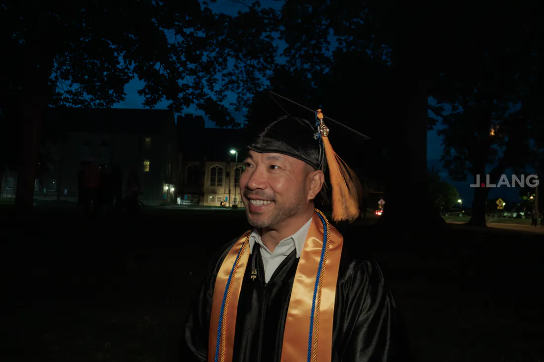 Smiling graduate in cap, tassel and gold stole on a campus lawn at dusk