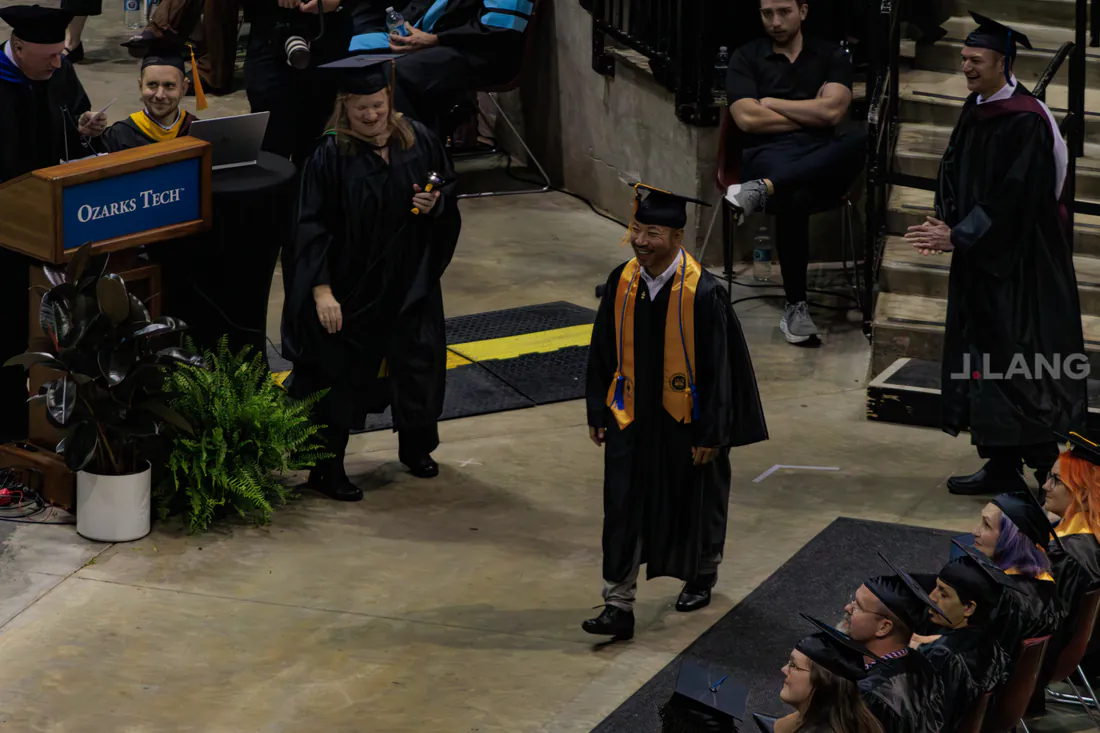 Smiling graduate in a gold stole crosses the commencement stage as robed faculty look on