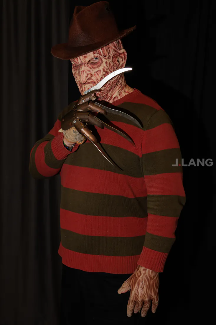 Cosplayer in a scarred mask, fedora and red and green striped sweater raising a bladed glove