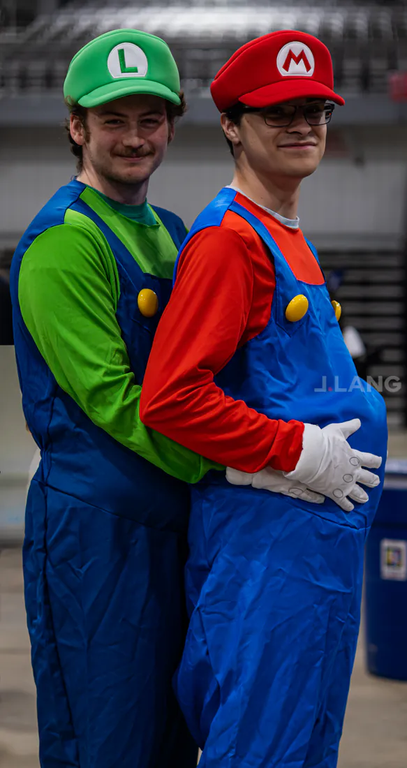 Smiling cosplayers dressed as Luigi and Mario pose together, Luigi hugging Mario from behind
