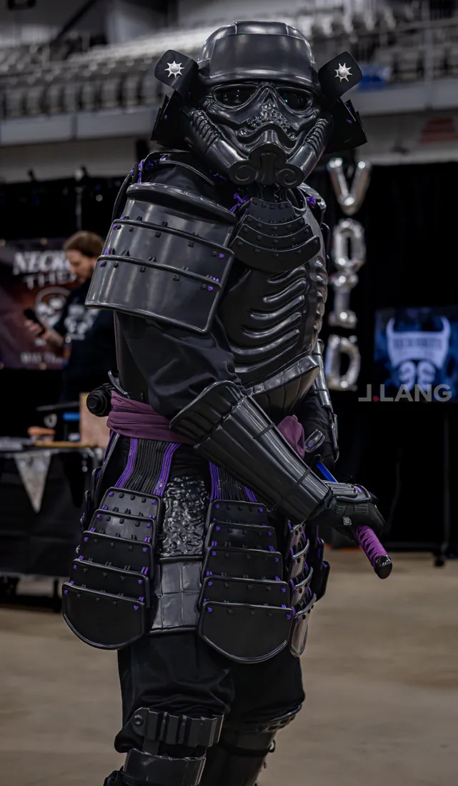 Cosplayer in black samurai armor and a stormtrooper-style helmet grips a sword in a convention hall