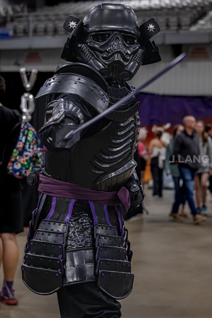 Black-armored samurai stormtrooper cosplayer holds a sword raised diagonally on a busy convention floor