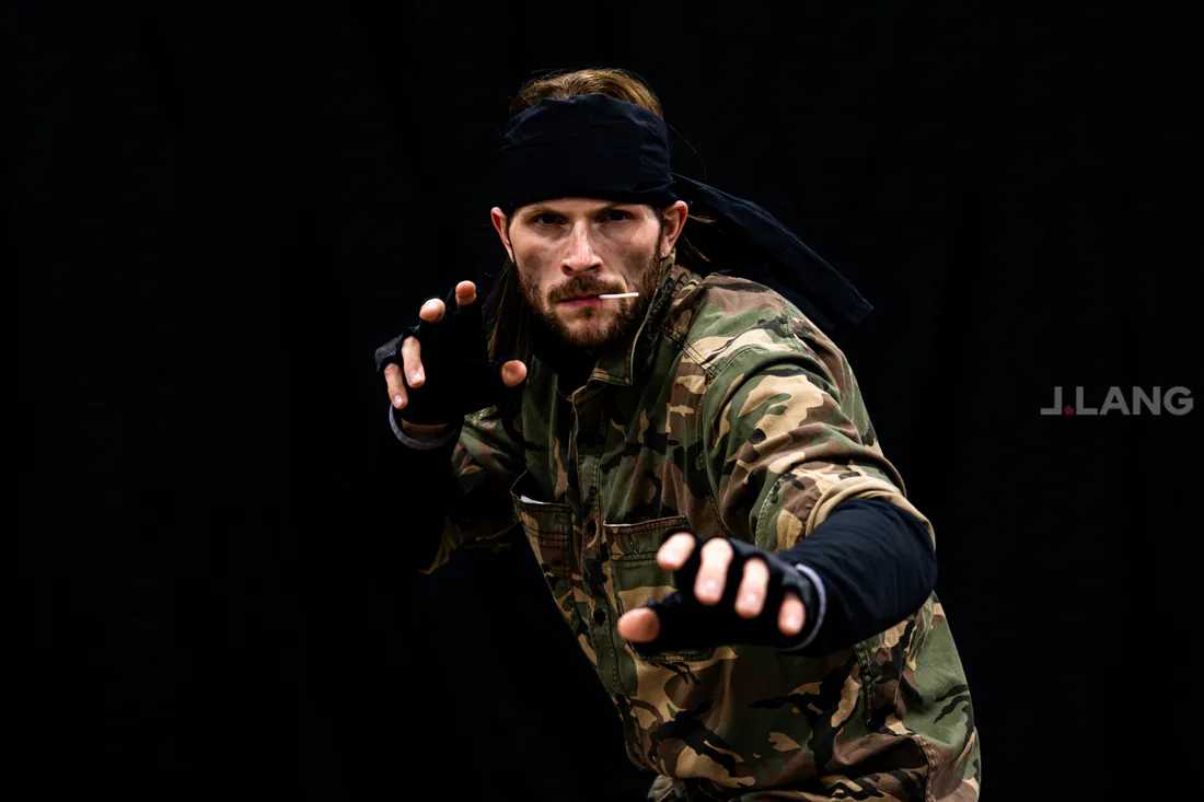 Bearded cosplayer in a camo jacket, black bandana and fingerless gloves takes a fighting stance