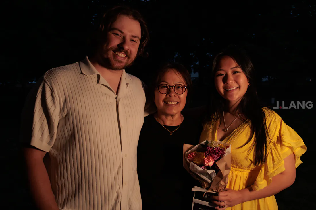 A bearded man and two women smile in warm light, one in yellow holding a bouquet