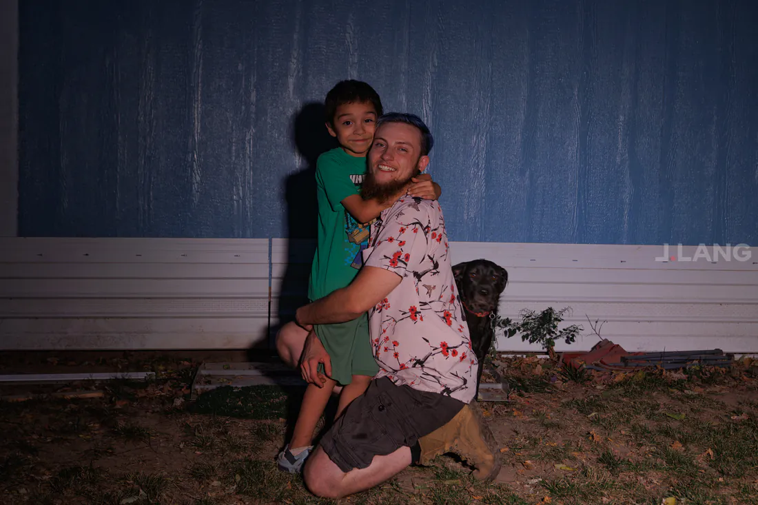 A kneeling bearded man and a young boy hug in a yard, a black dog nearby