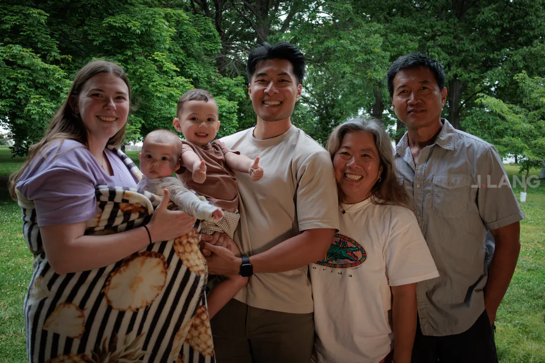 A smiling family in a park, a toddler giving two thumbs up beside a baby