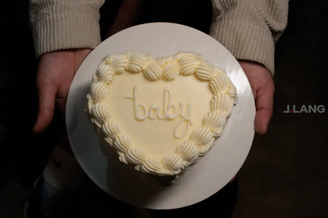 Hands hold a small heart-shaped cake with piped frosting and the word baby