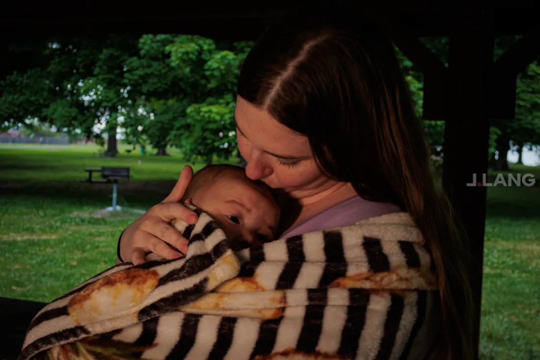 A woman kisses the head of a baby in a striped blanket, green park behind