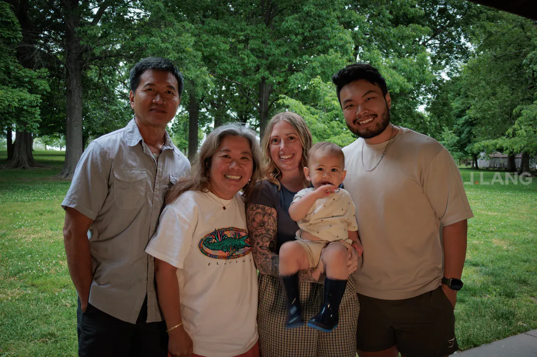 Four smiling adults pose in a park, a blonde woman holding a baby in rain boots