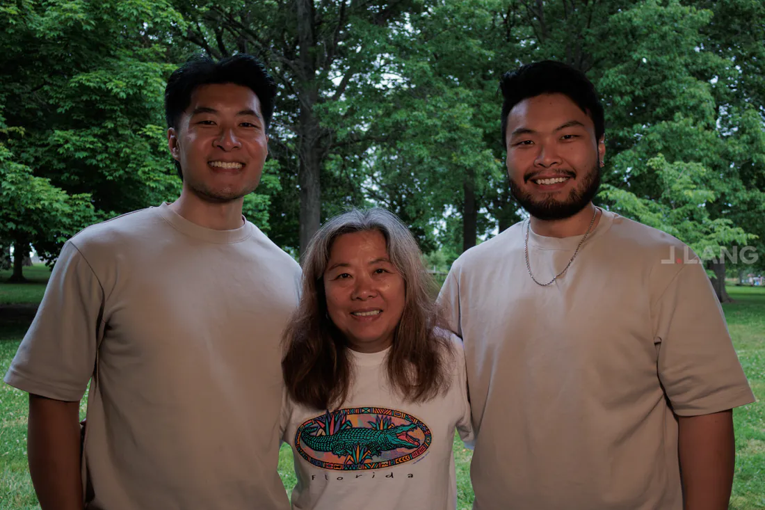 Two young men in matching tan shirts flank a smiling older woman under park trees