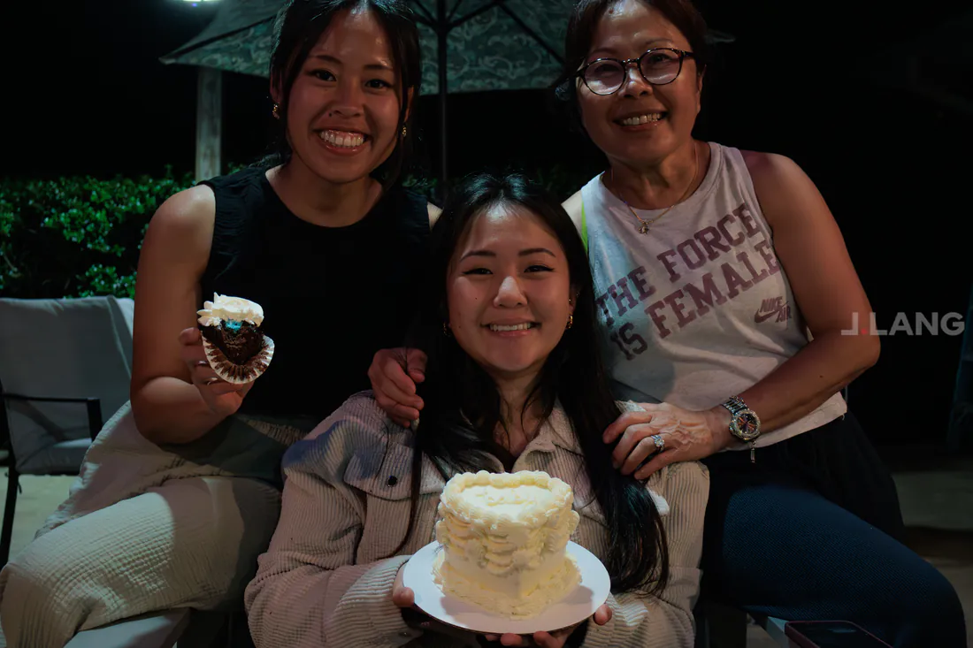 Three smiling women at night, one holding a heart cake, another a blue-filled cupcake