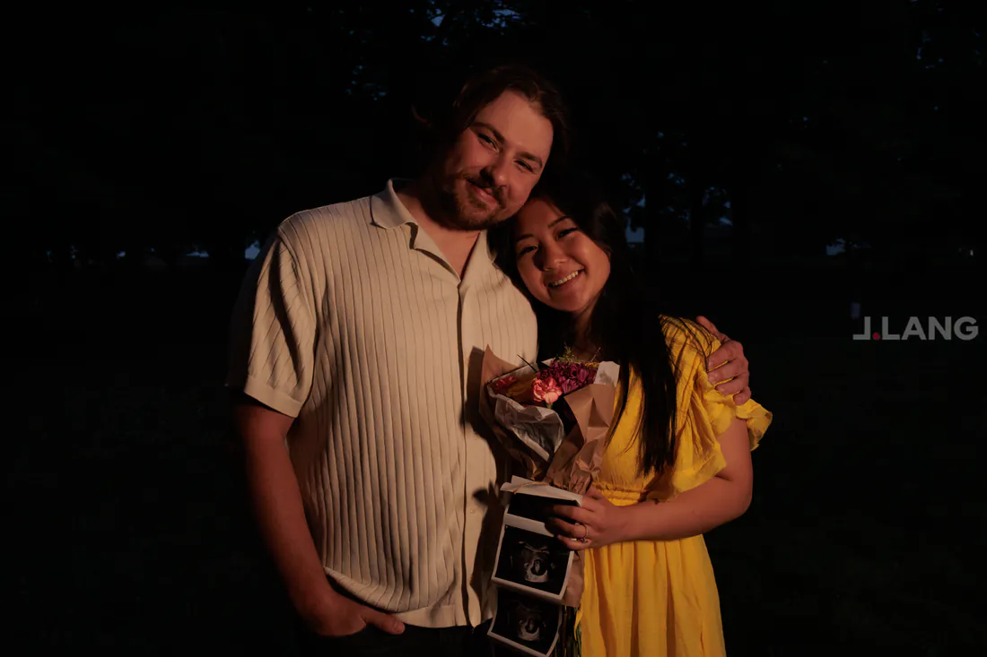 A couple leans together against a dark background, the woman in yellow holding flowers and ultrasound prints