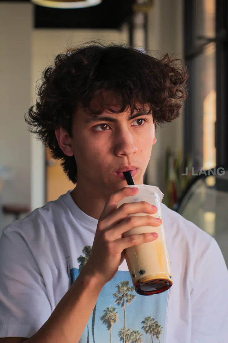 Curly-haired teen sipping a milk tea with boba through a straw, gazing to the side