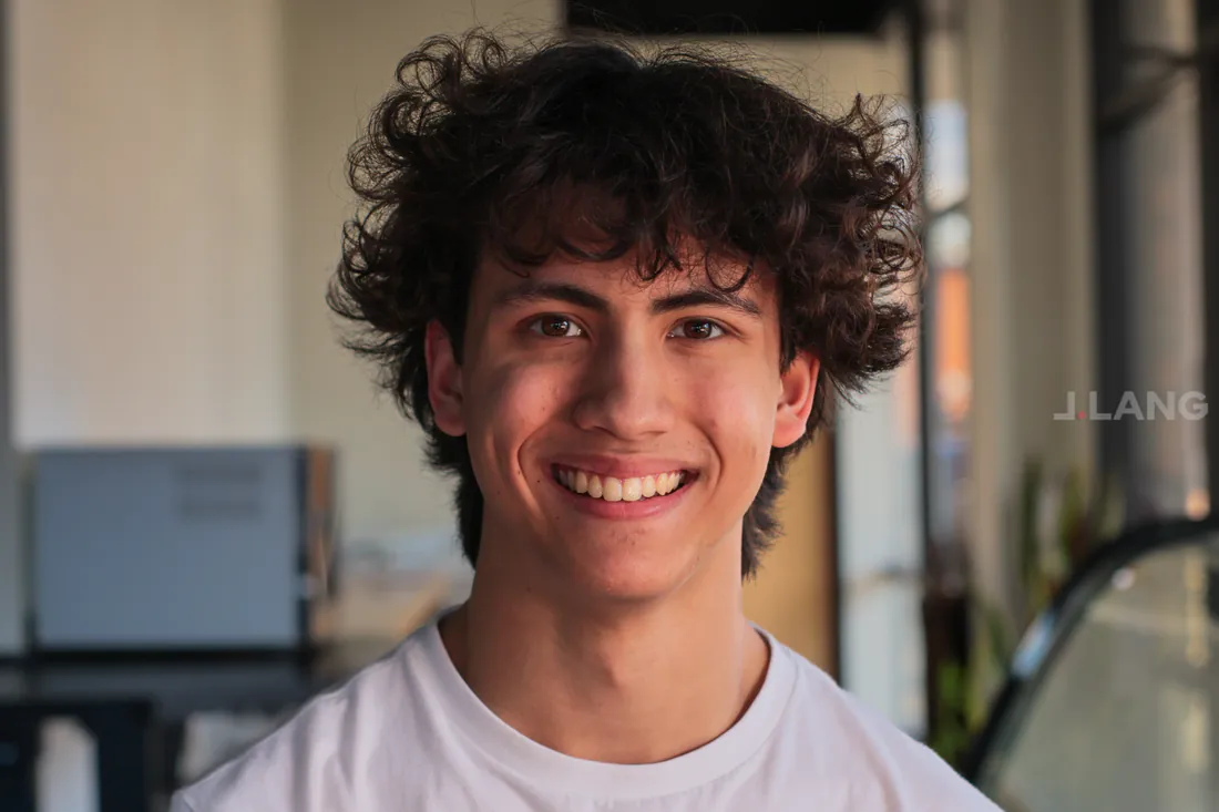 Teen with curly dark hair and a white T-shirt smiling broadly in a sunlit cafe