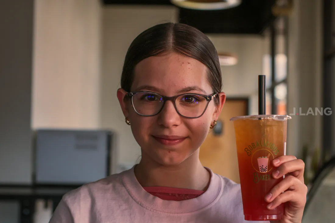 Girl in glasses and a pink T-shirt smiles, holding up a layered iced tea