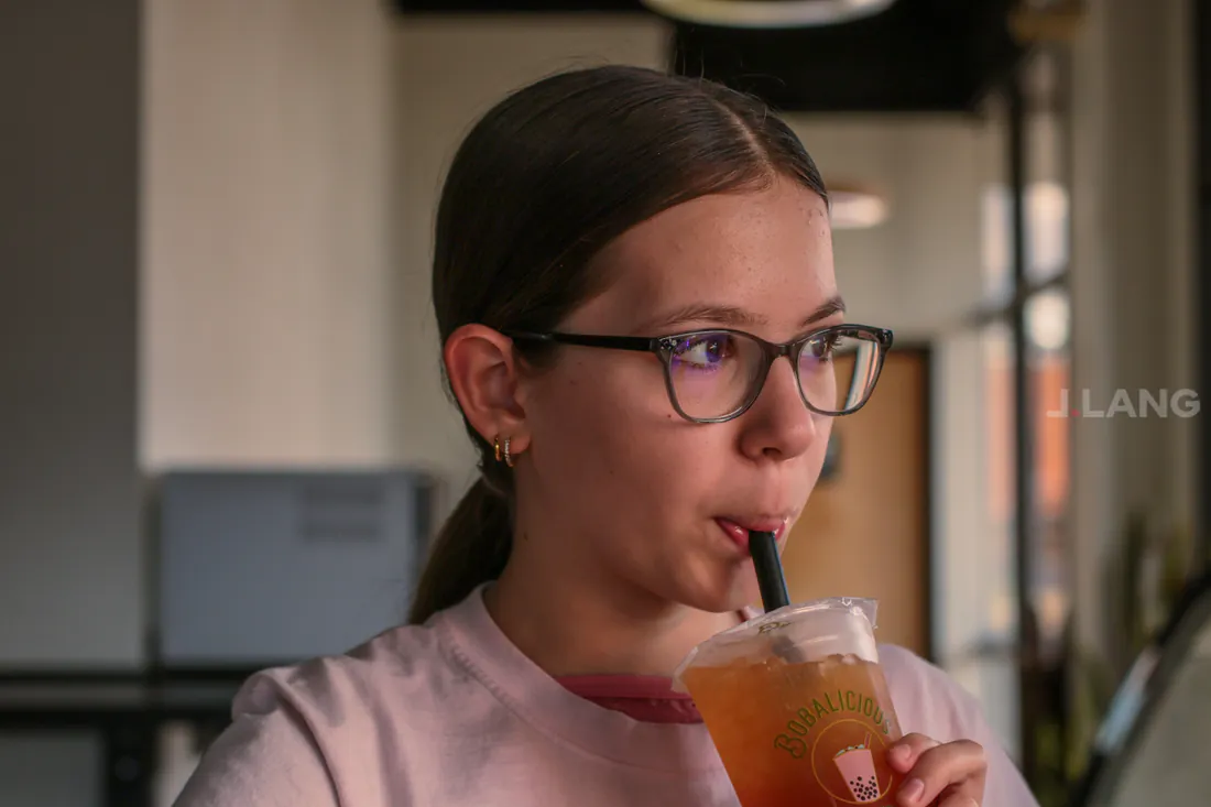Girl in glasses with a ponytail sips iced tea through a straw, looking to the right