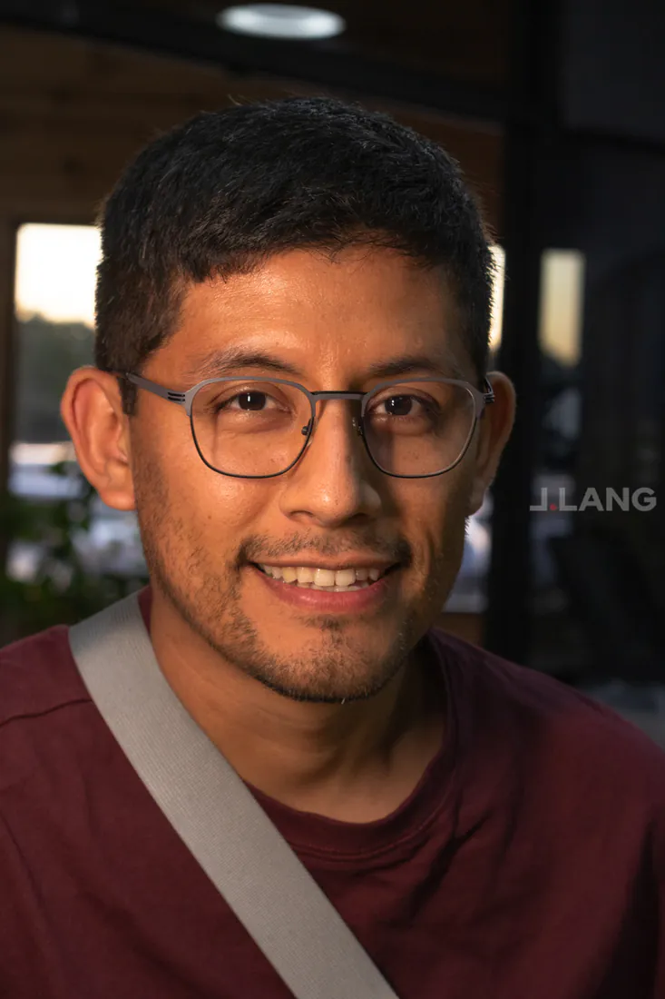 Smiling man in thin metal glasses and a maroon shirt, warm light behind him