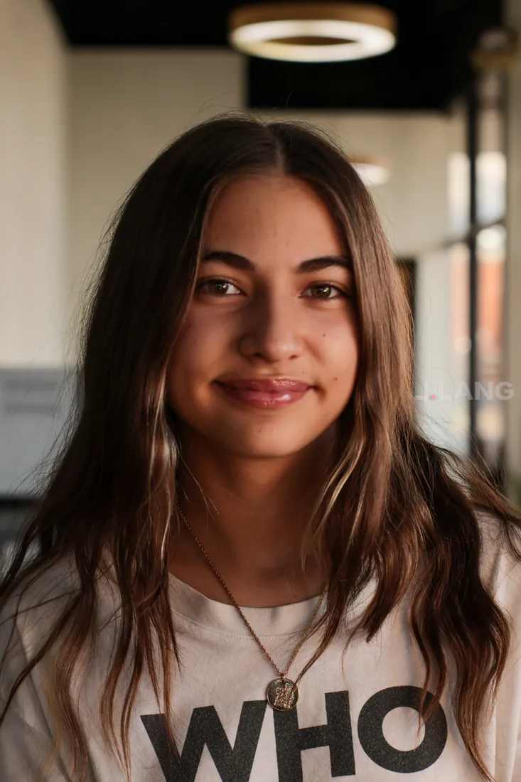 Teen girl with long wavy brown hair and a pendant necklace smiling softly at the camera