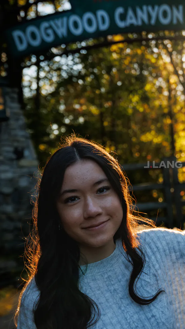 Backlit young woman smiles beneath a large entrance sign, autumn trees glowing behind her