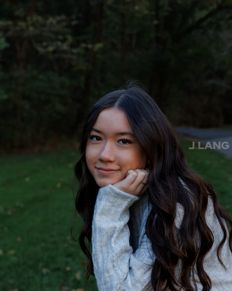 Smiling young woman with long wavy hair rests her chin on her hand before dark woods
