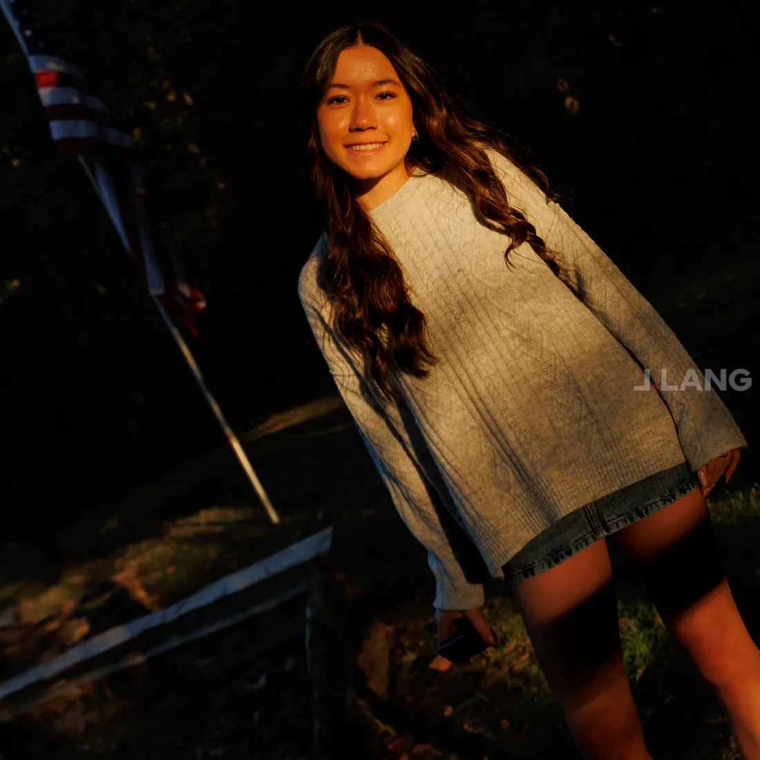 Smiling young woman in a knit sweater stands in low golden sun near an American flag