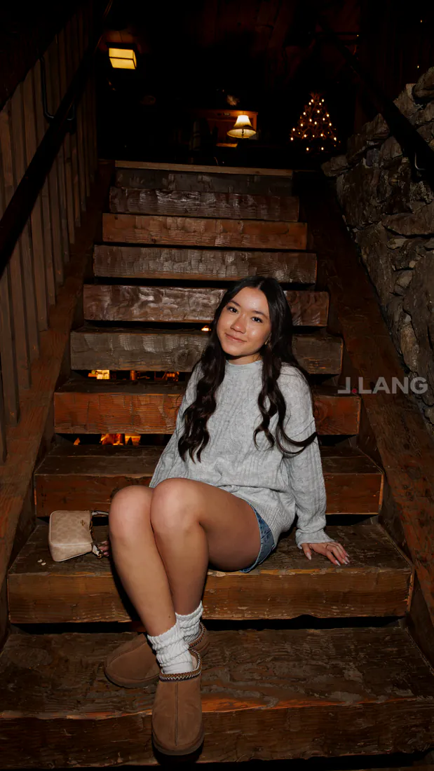Young woman in a sweater and shorts sits smiling on rustic wooden stairs indoors