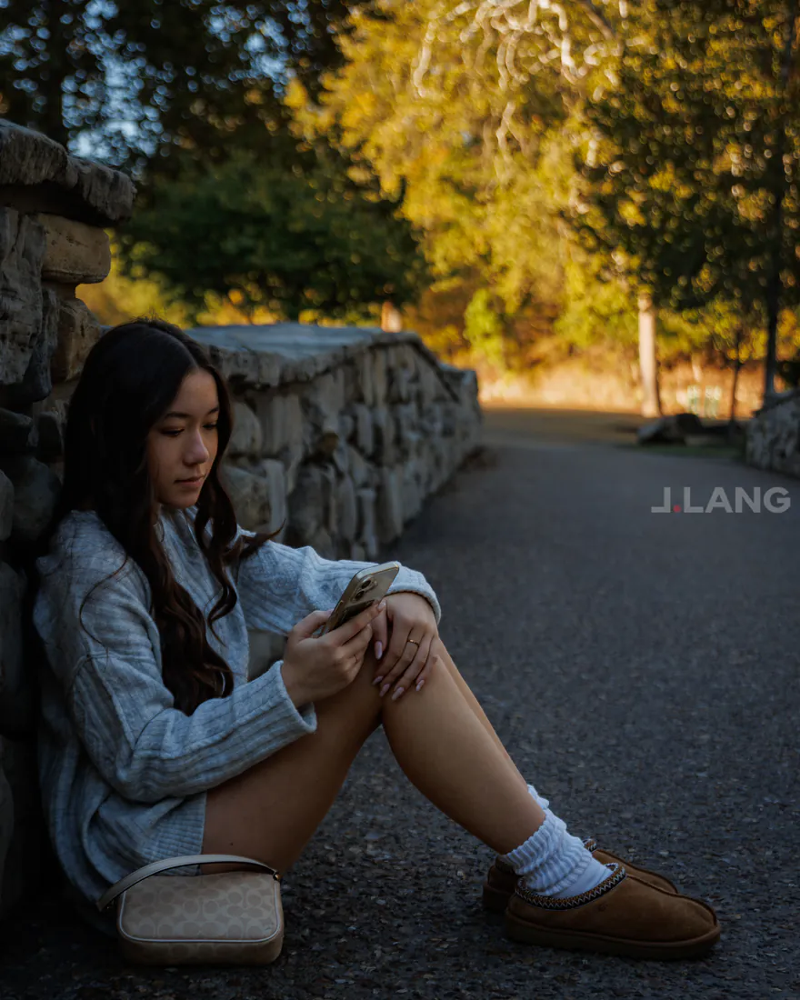 Young woman sits on a paved path against a stone wall, looking at her phone
