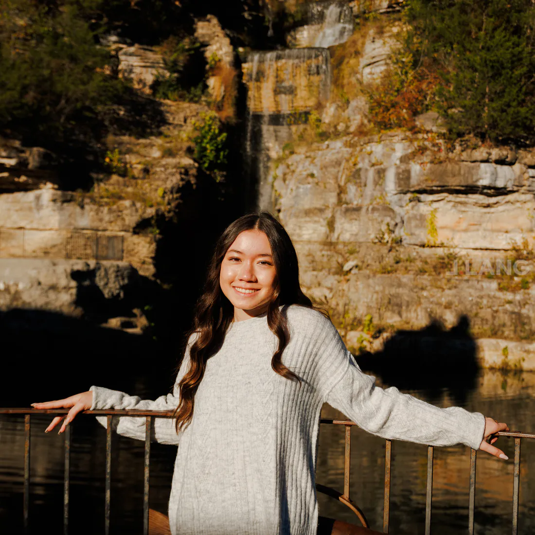 Smiling young woman rests her arms along a railing in front of a cliffside waterfall