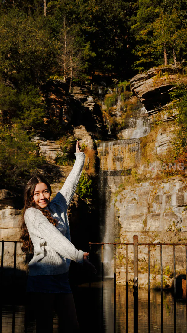 Smiling young woman points up toward a waterfall spilling over a tall rocky cliff