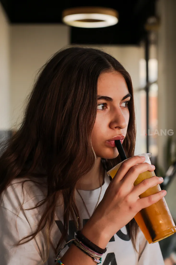 Young woman with long brown hair glances aside while sipping an orange drink in a cafe