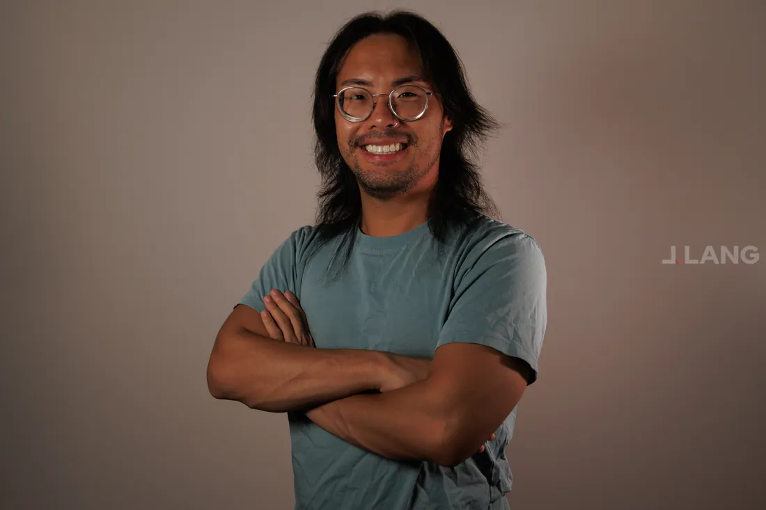 A smiling man in round glasses and a teal T-shirt stands with arms crossed