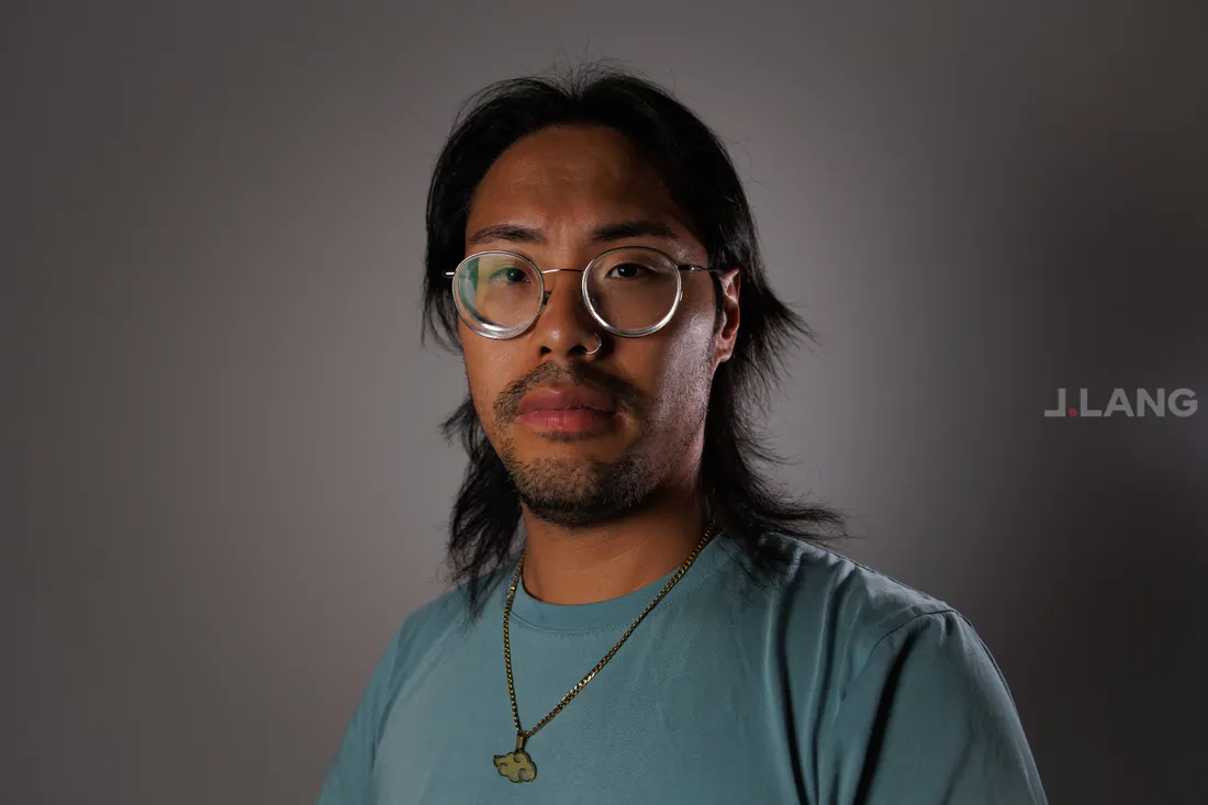 A serious man with long dark hair, round glasses and a gold pendant necklace