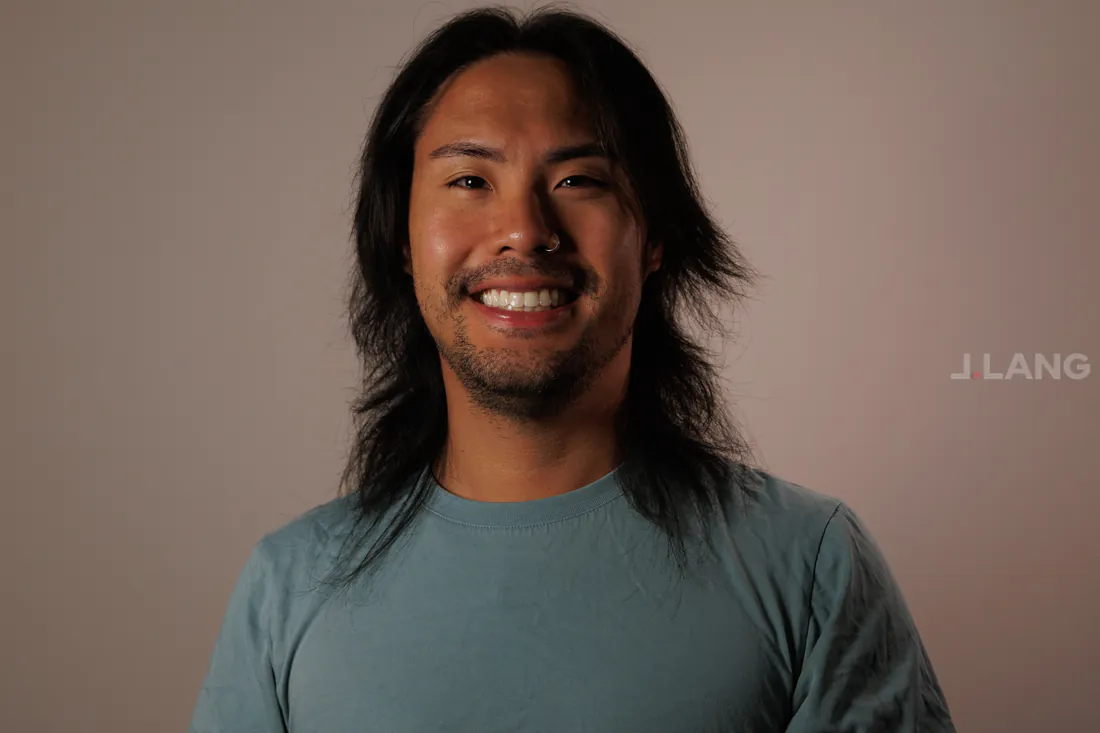 A smiling man with long dark hair and a nose ring against a plain studio backdrop